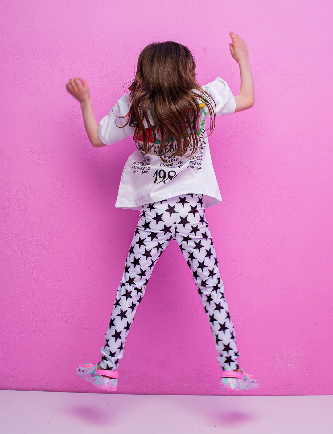 A child facing a pink wall jumping in the air with their arms up wearing white leggings with black stars 