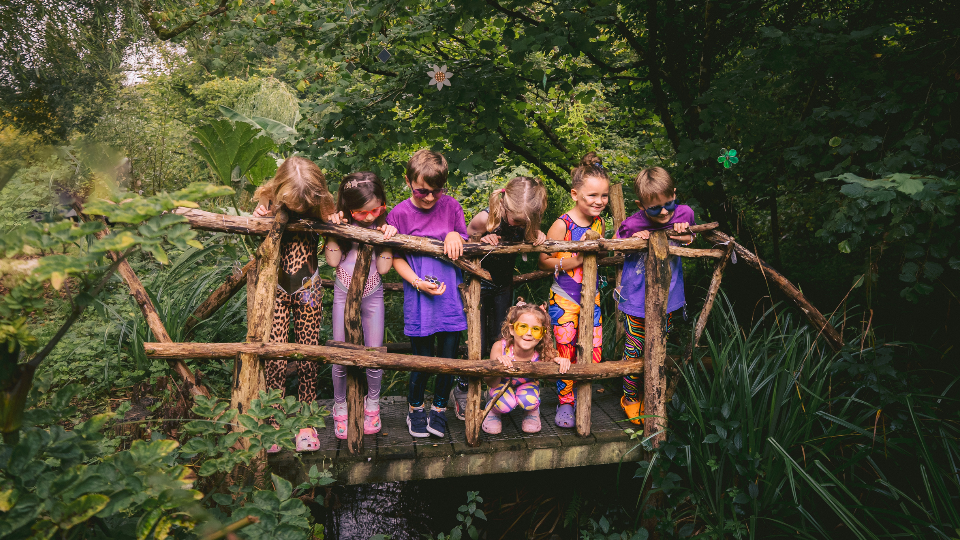 Children in colorful costumes standing on a wooden bridge in a forest setting
